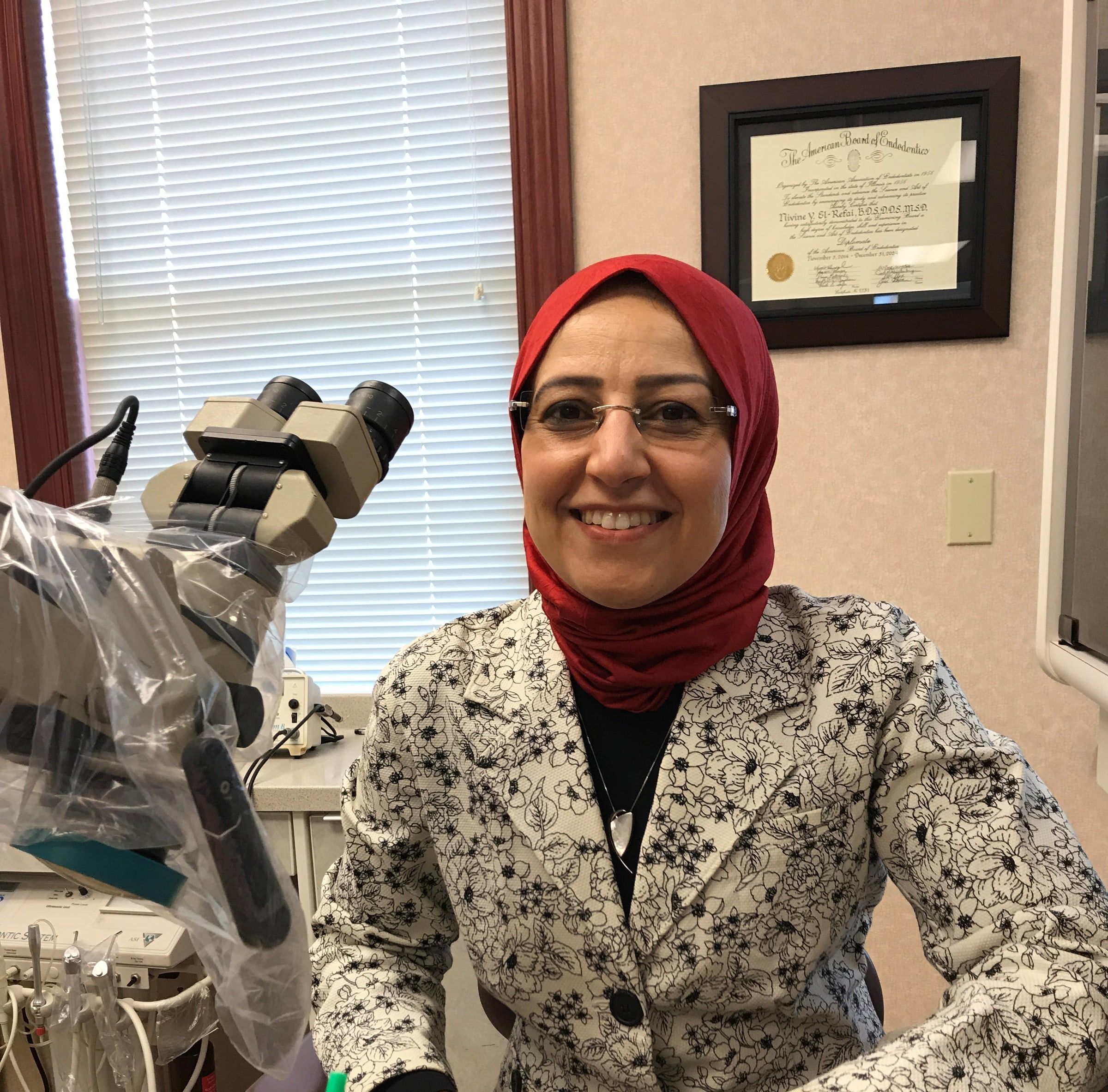 Dr. El-Refai smiling beside her surgical microscope, with her American Board of Endodontics certificate on the wall behind her